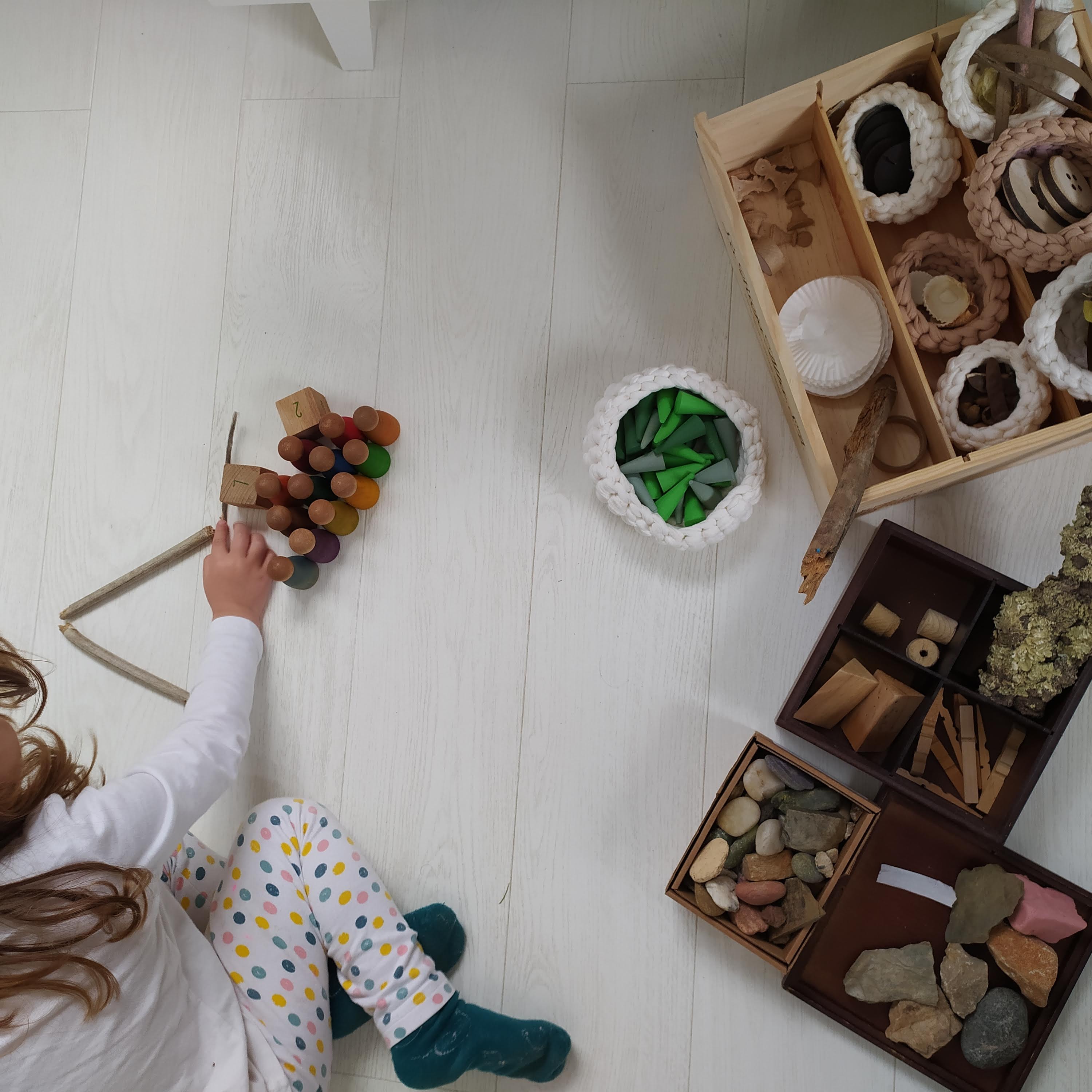 Pedres, botons de fusta, pals, nins, cons, retalls de fusta, escorça d'arbre... elements naturals classificats en cistells i una nena jugant